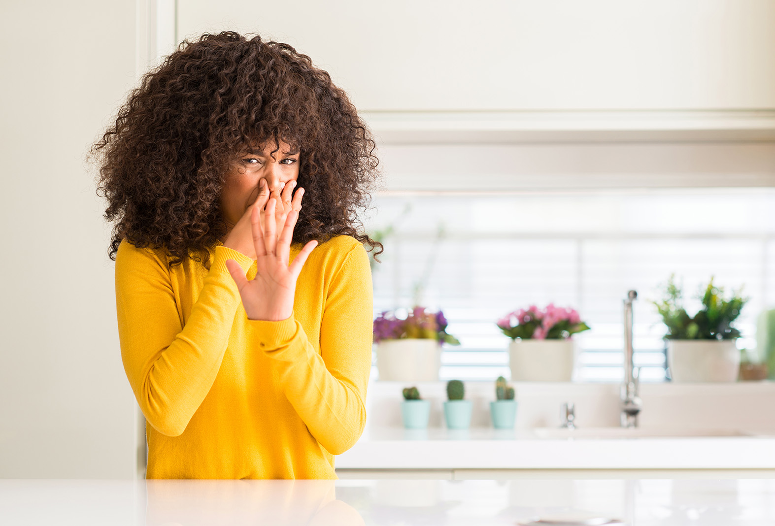 African american woman wearing yellow sweater at kitchen smelling something stinky and disgusting, intolerable smell, holding breath with fingers on nose. Bad smells concept. Woman holding her nose because of mold odors